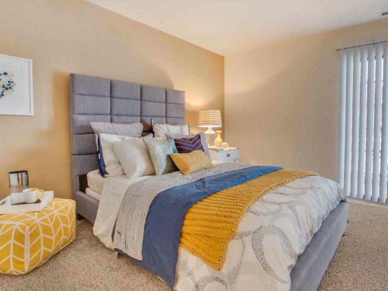 This image shows a master bedroom with modern lighting fixtures. It offers spacious floor plans with ample closet space and extra storage. It also features a multicolor blanket and pillows.
